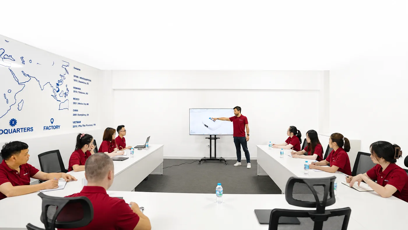 Casadevall staff seated around a U-shaped table in a briefing room, a colleague presenting at a screen beside a wall map marking the group’s headquarters and factories.